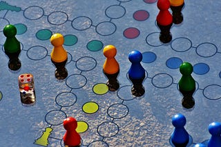 Overhead view of a multiplayer board game in progress, showing colorful game pieces in green, yellow, orange, blue, and red positioned on a blue game board with circular spaces and pathways. A die is visible on the left side, and the pieces cast shadows on the board, illustrating the original form of multiplayer collaboration and competition that preceded digital gaming.