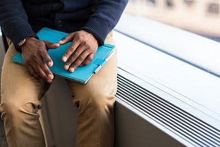 A Black job seeker’s hands rest on their lap on top of a tablet in a blue cover. They seem to be positioned as if the photo was taken while the person was explaining something.