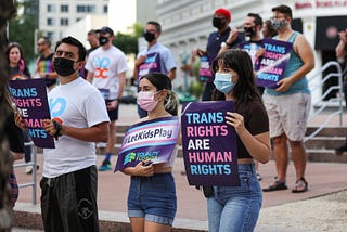 Masked protestors of different genders and races stand with signs that read “Trans Rights are Human Rights” and “#LetKidsPlay Equality Florida”