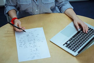 A woman has both hands over a wooden table. One hand is over a laptop, while the other holds a pen and is drafting UI screens