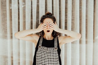 A woman is covering her eyes with her hands. She is wearing a black top and black checked overalls.