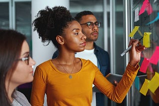 A woman writes on pink and yellow post-it notes on a glass, while two people watch.