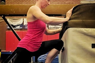 The author wears a pink tank top and black leggings, lunging forward in a calf stretch next to a balance beam