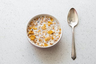 A bowl of cereal with milk and a silver spoon rest on a table top