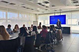 A professional corporate training session for Touchpoint Strategies by Knowcrunch is taking place in a modern conference room, where a presenter stands near a screen displaying a welcome message while participants engage in discussion around a large table.