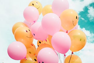 Photo showing a collection of balloons with a sky background. Half of the balloons are yellow with a happy smilie face, the other half are pink with a sad smilie face.