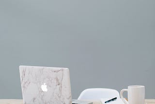 An open MacBook and notebook sit on a wood desk next to a cup of coffee, indicating unfinished work. The chair behind them is empty.