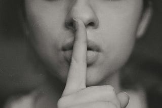 Black and white photo of a woman shushing with index finger of lips.