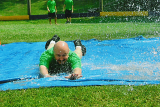 It’s a sunny, hot summer day, and a bald man is sliding face-forward toward the camera on a homemade slip-n-slide in a beautiful, green field. He is in a green shirt and you can see his while legs and black shoes as he’s almost to the end of the watered down, blue tarp. To young men in are in the background watching him (with matching green shirts and dark shorts) waiting their turn to “slide.”