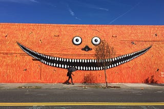 Monster Mural on a wall in Portland Oregon. Big wide smile against a reddish orange background. Person walking past in the foreground carrying a black umbrella.