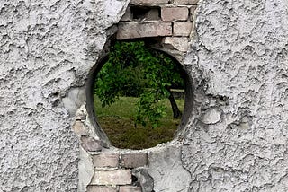 A grey brick and cement wall with a round hole in the centre through which some grass and a tree can be seen