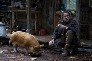 Robin (played by Nicolas Cage) sits on the porch of his cabin holding a piece of pie. His brown pig is next to him and eating leftovers out of a skillet.