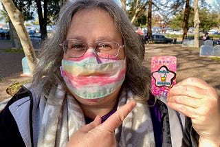 Michele Mashburn, a white woman with graying hair and glasses, wearing a mask pointing at a “Suck it Ableism” pin. There are trees behind her.
