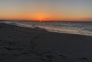 The sun rises out of the ocean, it looks like a pink peach appearing out of the dark-velvety-blue, calm ocean.