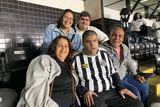 Foto do torcedor santista Matheus e sua família nas arquibancadas da Vila Belmiro, estádio do Santos. Na primeira fileira, Matheus está sentado entre seu pai e sua mãe, com outros dois familiares do torcedor na fileira de trás.