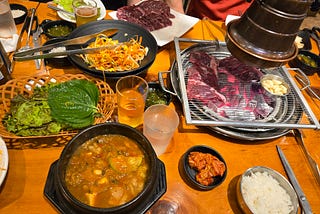 Table with soup and vegetable side dishes, and a grill cooking pieces of meat.
