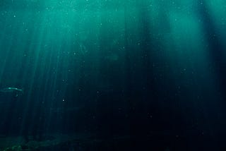 fundo do mar, com raios de luz atravessando a imagem em linhas verticais. As águas fazem um degradê de verde azulado até completamente preto.