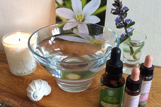 A glass bowl filled with water, surrounded by essences, a shell, a lit candle, a flower card, and a sprig of blue flowers