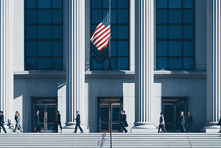 Professionals walking up the steps of a federal government building with tall columns and an American flag, representing the U.S. contracting and business sector. Discover how small federal contractors can create lasting growth through operational flywheels that compound performance, trust, and capability.