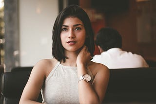 Brown-eyed woman with tan skin and black bob is seated on a black couch in a public place. She looks toward the camera with a neutral but curious expression.