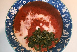 Una sopa roja en un cuenco azul. Hay una espiral blanca de leche de coco y una montañita de semillas de calabaza.