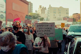 A group of people protesting for abortion rights and reproductive justice. They are wearing face masks and many are holding protest signs. A more prominent sign reads: My Body My Choice My Future. Safe abortion saves lives.