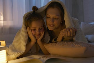 A girl and a woman are cozily nestled under a blanket, enjoying a book together by the warm glow of a bedside lamp. The adult is reading aloud, engrossed in the story, while the younger one listens intently with a look of delight. The atmosphere is calm and intimate, perfect for a bedtime story.