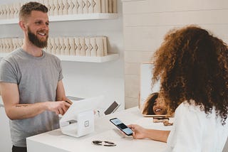 A woman purchasing a product using her smartphone as a payment method with a man smiling as he sells the product.