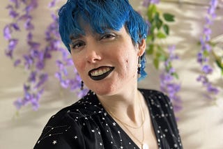 Image of Tori, a white femme with blue hair and black lipstick. She has earrings with crystal beads in white, purple, grey and black. She has dark eyeshadow and white graphic eyeliner shaped like stars. She is wearing a black dress with small white constellations on it. She is absolutely beaming and only has one acne scar on her left cheek. She is wearing a layered gold necklace with a real flower in it.