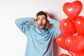 Boyfriend grabs his head with his hands in a panic about Valentine’s Day, heart balloons next to him, an alarmed look on his face.