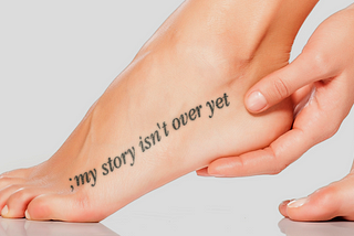 Image of a woman’s foot, with her heel cupped in her fingers. A semicolon and these words — my story isn’t over yet — are tattooed from her pinky toe to her ankle.