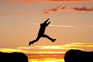A silhoutte jumping between two cliff edges in front of a setting sun