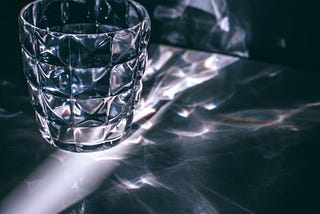 A multifaceted glass sits on a table in a dark room, caught in a shaft of light from a slightly open door, scattering reflected light on the table. Photo by Maria Orlova: https://www.pexels.com/photo/glass-with-refraction-pattern-in-darkness-4946525/