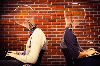 Two people with light bulbs for heads in front of a brick wall, typing on tablet and phone.