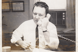 Classic 1960s man working the phones for the DNC in November 1961. He has on a white work shirt, tie, and is using a landline.