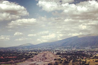 An Aztec pyramid is in the centre of the photo, sorrounded by scarce vegetation, with some mountains in the distance.
