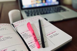 An open diary with a list of highlighted writing goals laid in front of a MacBook