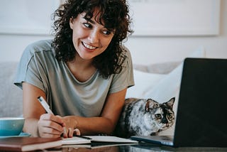 Person using laptop takes notes in a journal. A cat rests beside them, also staring intently at the screen.