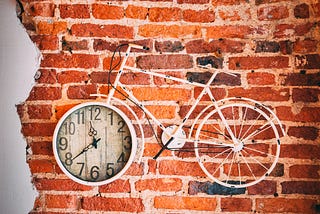 A white bicycle hangs in front of a brick wall, the front wheel is a clock