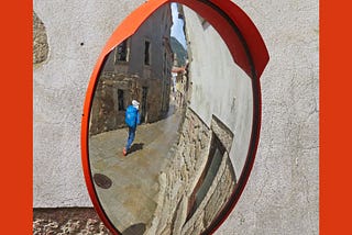 Backpacker with a bright blue backpack’s reflection in a round mirror, is seen walking down a medieval street, away from the camera. There is an orange sun visor on the mirror and that is reflected in an orange frame around the photo.