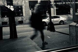 Black and white blurry shot of a stranger walking on the street while a car passes by
