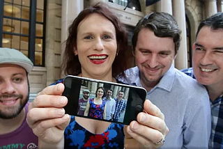 Four people stand in front of the Apple Store in Brisbane’s Queen St, one holds up a cellphone which displays an image of the four.