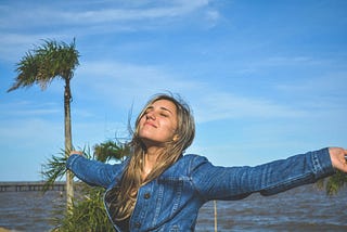 woman in blue jeans jacket, eyes closed, facing sun in breeze near water