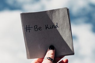 A hand holding a napkin with the hashtag “Be Kind” written on it