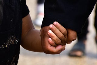 child holding adult’s hand, both brown-skinned wearing black clothes