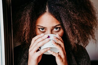 A woman sips coffee. She is holding the coffee cup with two hands, looking directly (and intensely) at the viewer.