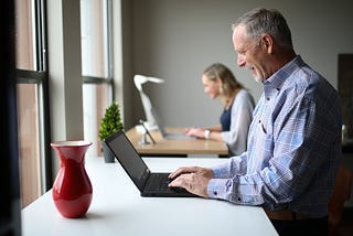Stock image of two people at standing desks working on laptops. The person in the foreground is an older man. The person in the background is a younger woman.