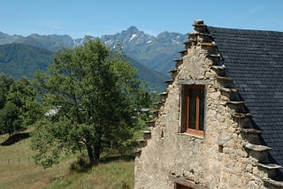 A stone barn with stepped gables and pastures with rocky mountains in the background.