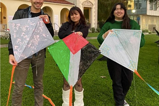 Three students stand on top of a hill, each holding a white kite. The kite on the right has “Free Palestine” written multiple times in light pink. The middle kite has the Palestine flag drawn on it — a red square at the top, with a green stripe on the right and a black stripe on the left. The kite on the left has “ASA” written in green, with green flowers on the border.