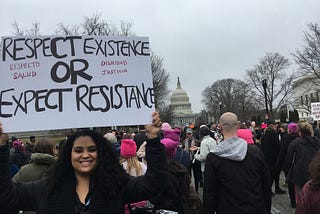 Photo of Emmeline Rodriguez holding a sign saying ‘Respect Existence or Expect Resistance’ in English with smaller red lettering reading ‘Respecto Salud / Dignidad Justicia’ in spanish. She is on the street joined by other protesters near the Capitol building in Washington D.C. All people in the photo wear warm clothing, including Emmeline who wears a black jacket and black scarf.
